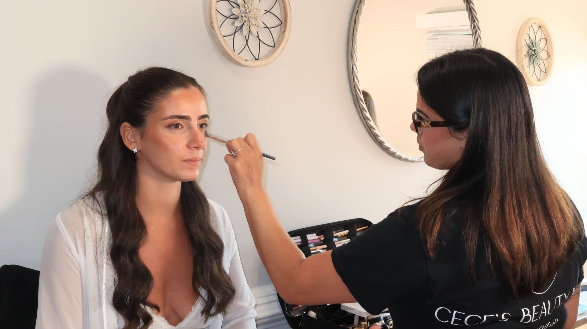 A makeup artist applying makeup to a woman, with beauty products and brushes visible in the background.