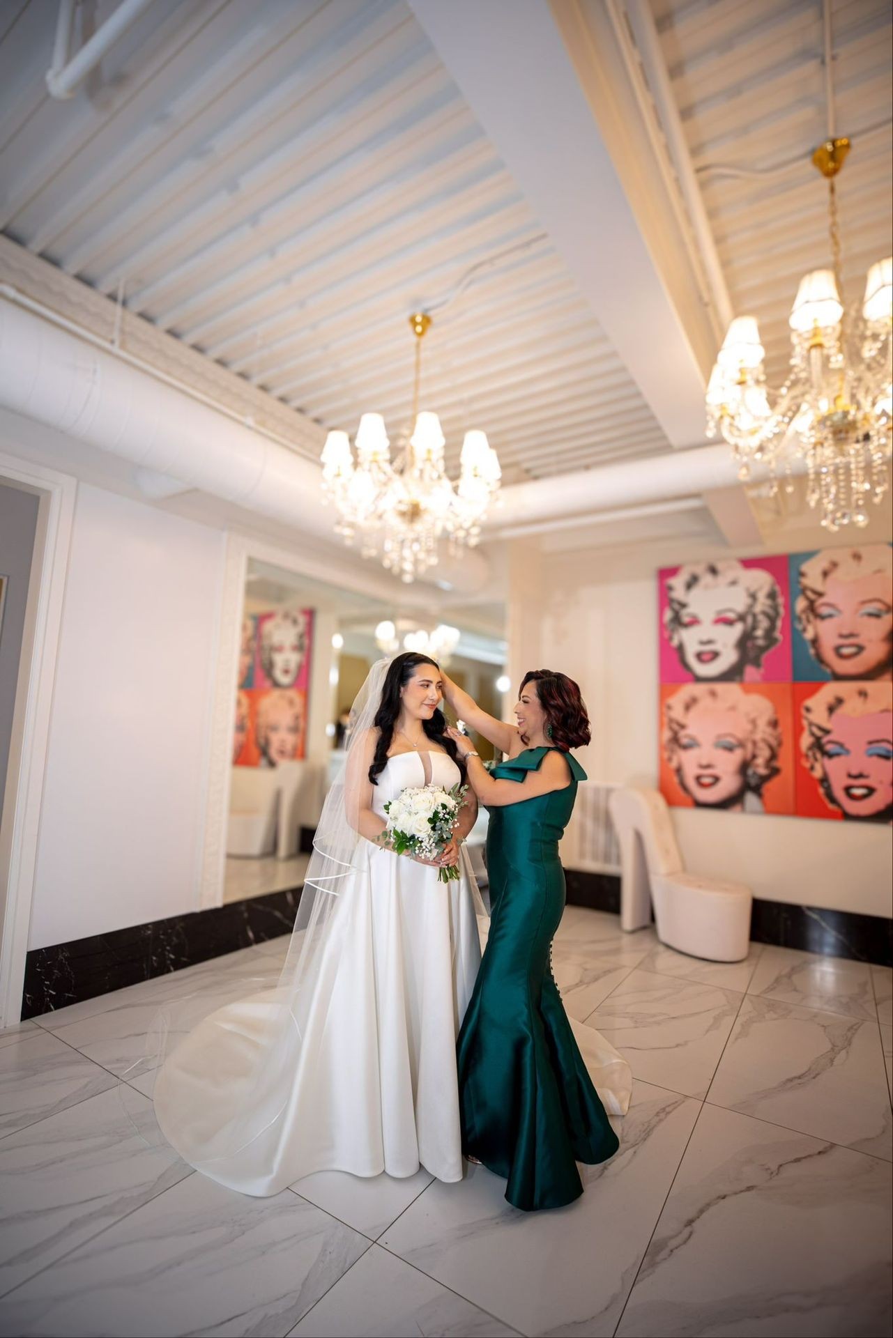 Bridesmaid adjusts bride’s veil in elegant room with chandeliers and pop art portraits.