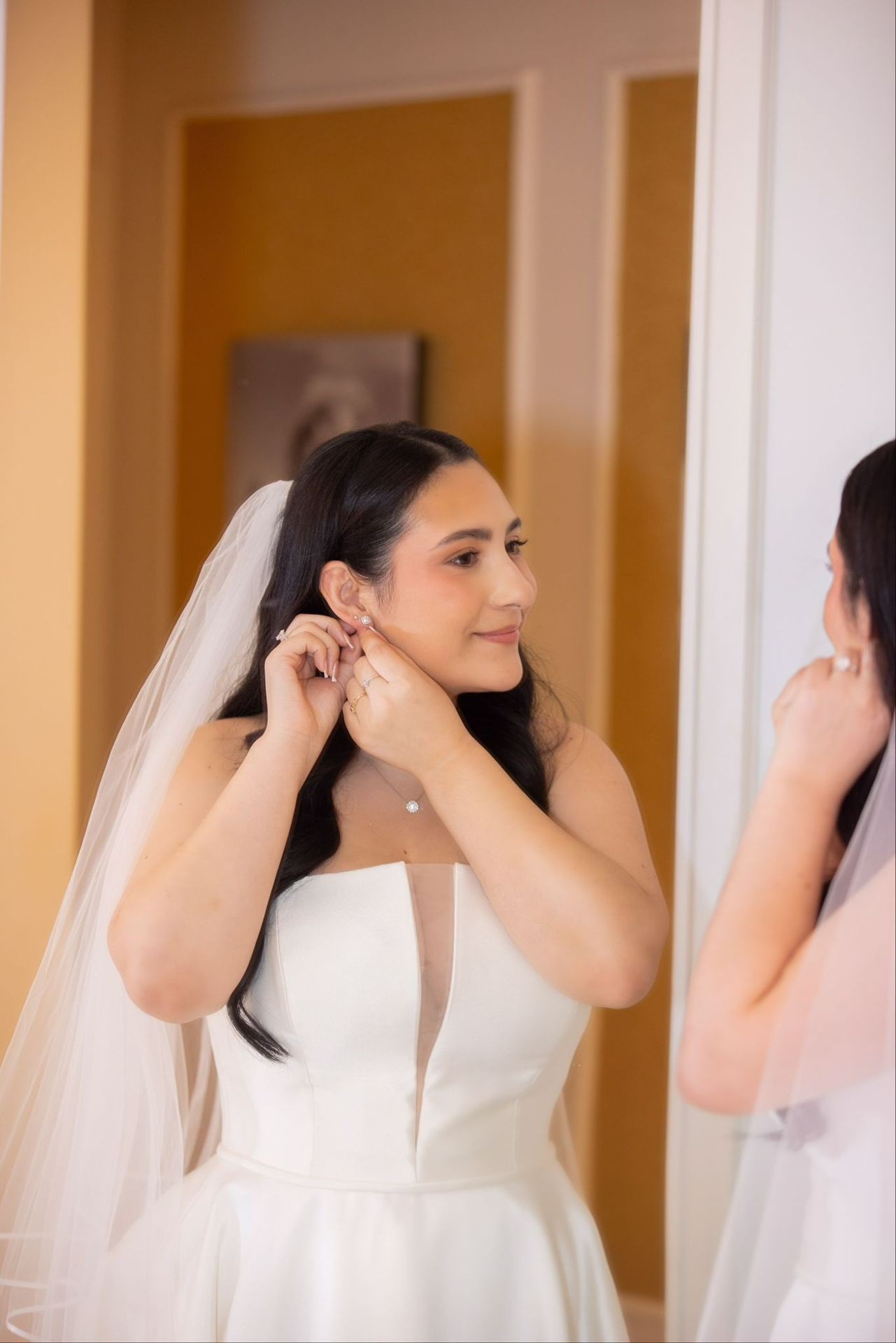 Bride in white dress and veil adjusting earrings in front of mirror.