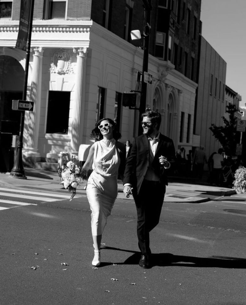 Black and white photo of a couple in formal attire holding hands and walking on a city street.