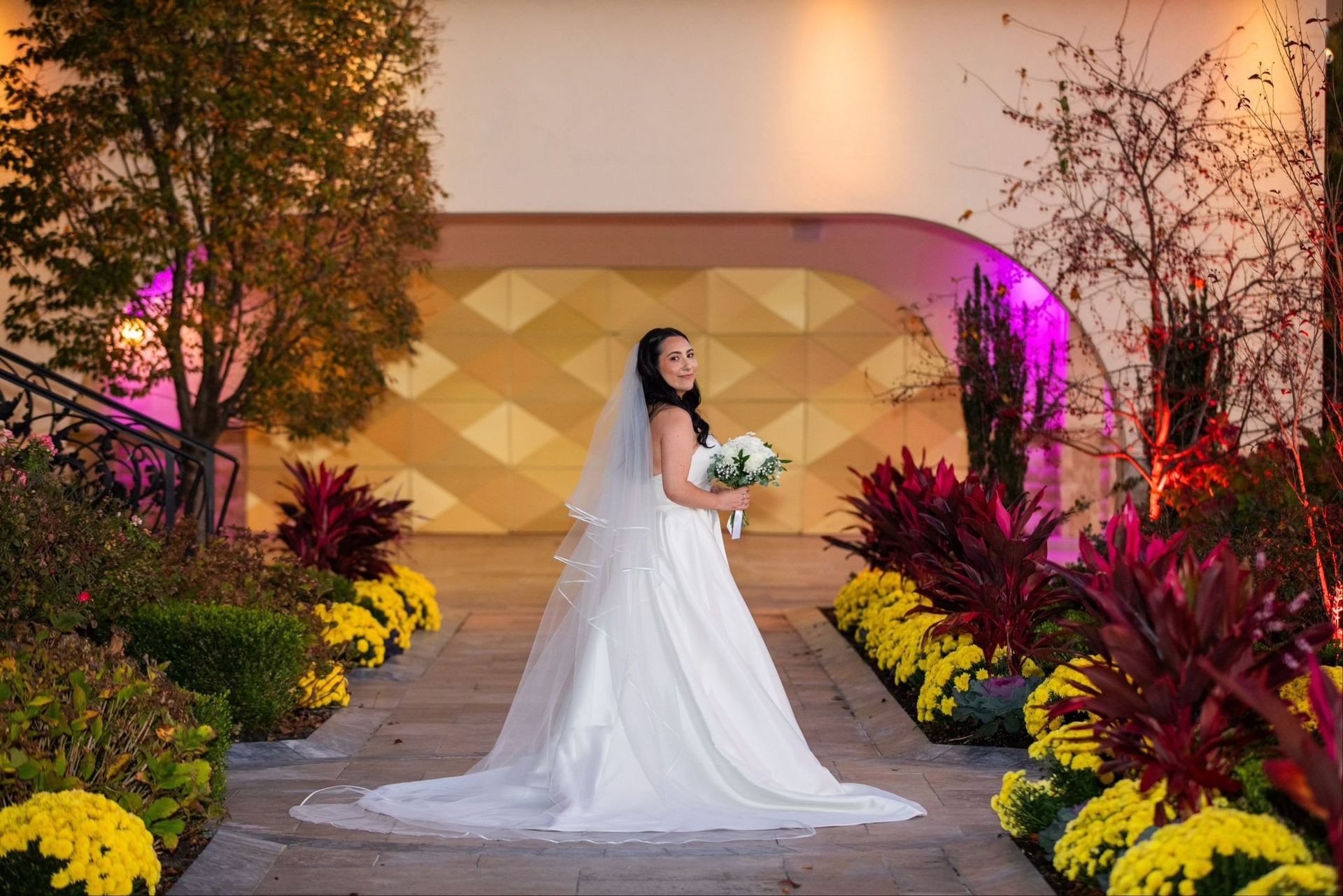 Bride in a white gown holding a bouquet, standing on a flower-lined pathway with decorative lighting.